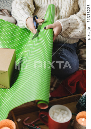 Young woman sits on plaid in cozy knitted woolen white sweater, socks and wraps Christmas gift in polka dot wrapping paper. Wooden tray with mug of cocoa with marshmallows, toy tree, candle, straws. Young woman sits on plaid in cozy knitted woolen white sweater, socks and wraps Christmas gift in polka dot wrapping paper. Wooden tray with mug of cocoa with marshmallows, toy tree, candle, straws. 71338228