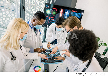 Top angle view of multiethnical team of doctors in medical masks, examining CT scan together, discussing diagnosis and ways of successful treatment. Medical workers looking at scan at the hospital 71340237
