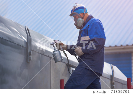 Industrial work. Priming of metal products from the compressor gun. A worker in overalls and a respirator paints the body of a truck trailer or a metal car. 71359701