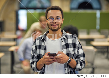 Portrait of a young happy mixed race man holding smartphone and looking at camera while working in the modern office, selective focus Portrait of a young happy mixed race man holding smartphone and looking at camera while working in the modern office, selective focus 71363114