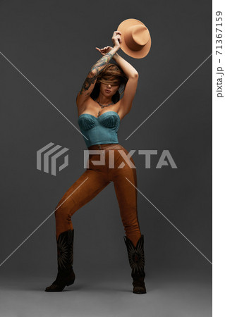 Fashion portrait of young brunette girl in blue western top and jeans with cowboy hat and boots in front of gray copy space studio background. Fashion portrait and lifestyle. 71367159