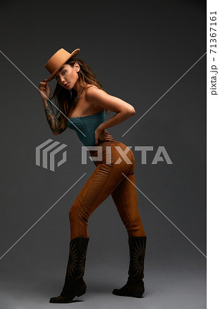 Fashion portrait of young brunette girl in blue western top and jeans with cowboy hat and boots in front of gray copy space studio background. Fashion portrait and lifestyle. 71367161