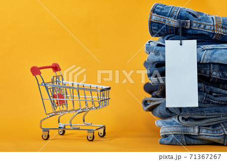 A stack of blue jeans with a white blank tag on a yellow background. Shopping trolley with multi-colored cubes. Sales word written on cubes. Sales consept 71367267