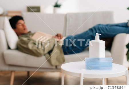 E-learning during covid-19. Black teen sleeping on sofa with textbook, selective focus of mask and antiseptic on table E-learning during covid-19. Black teen sleeping on sofa with textbook, selective focus of mask and antiseptic on table 71368844