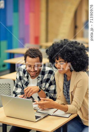 Two young happy diverse colleagues looking at laptop and discussing something while sitting at the desk in coworking space 71390976