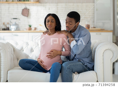 Worried black husband supporting pregnant wife with prenatal contractions at home 71394258