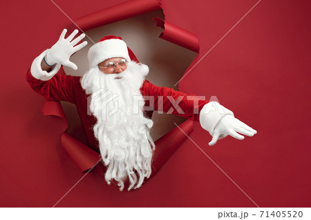 Santa man spread his arms in different directions and look emotionally through a paper hole. Bearded man in santa hat looking through hole on red paper 71405520