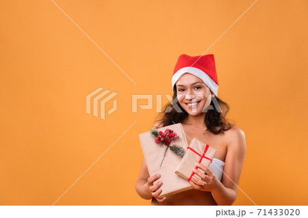 A young woman in a Santa Claus hat shows off Christmas presents and smiles. Portrait on a yellow background. Christmas concept. A young woman in a Santa Claus hat shows off Christmas presents and smiles. Portrait on a yellow background. Christmas concept. 71433020