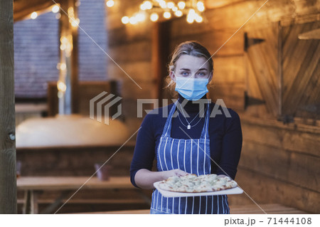 Woman waitress in apron and face mask holding fresh pizza on a board 71444108