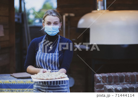 Woman waitress in apron and face mask holding fresh pizza on a board 71444114