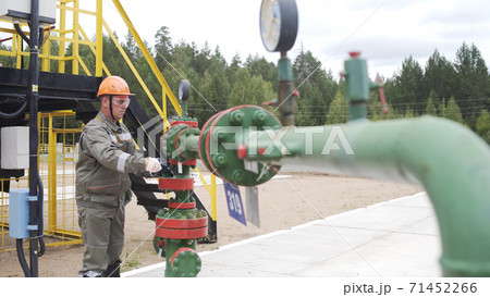 Oilman in orange helmet and protective glass opening or closing valve of wellhead. Injection tree is destined for injection wells sealing and their operating regime regulation. Oil industry Oilman in orange helmet and protective glass opening or closing valve of wellhead. Injection tree is destined for injection wells sealing and their operating regime regulation. Oil industry 71452266