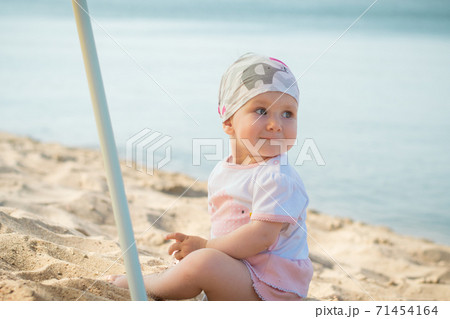 Little girl child in summer dress sits on beach beach or sea and lake and playing with yellow sand Little girl child in summer dress sits on beach beach or sea and lake and playing with yellow sand 71454164