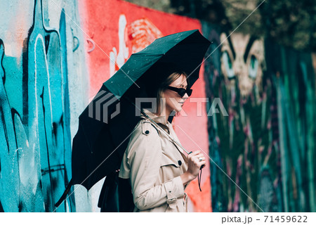 young stylish woman wearing long beige coat, white boots, black hat, umbrella and backpack posing through the city streets. Selective focus, grain 71459622
