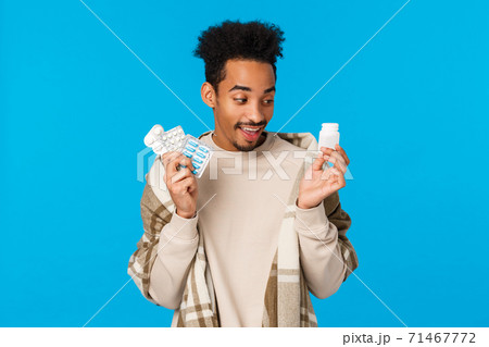 Funny handsome african man choosing what pill take first as got sick, staying home with cold or flu, holding pills from drugstore, use medication get better, taking care health, blue background Funny handsome african man choosing what pill take first as got sick, staying home with cold or flu, holding pills from drugstore, use medication get better, taking care health, blue background 71467772