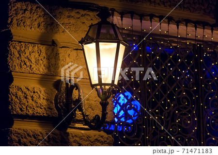 black wrought iron lantern with frosted glass shines with warm light on the facade of a historic building with rustication, in the background a gate with a garland on a dark night close-up, nobody. 71471183