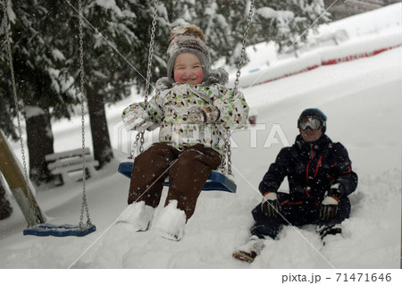 Little smiling child with red cheeks swinging on swing under snowfall and young father sitting on snow in snowy park 71471646