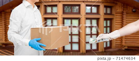 deliveryman hands in sterile gloves of courier cardboard box and hand holds out money to pay for food delivery during quarantine of social distance pandemic on background wooden building house. deliveryman hands in sterile gloves of courier cardboard box and hand holds out money to pay for food delivery during quarantine of social distance pandemic on background wooden building house. 71474634
