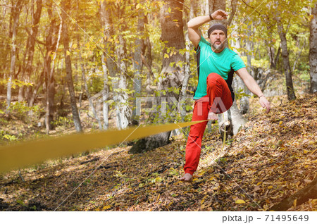 A bearded man in age balances while sitting on a taut slackline in the autumn forest. Outdoor Leisure 71495649