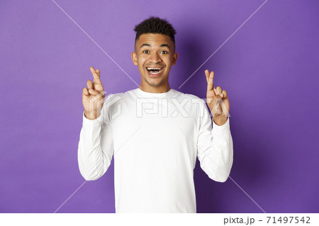 Image of handsome african-american young guy in white sweatshirt, looking hopeful and smiling with crossed fingers, making wish or waiting for good news, standing over purple background 71497542