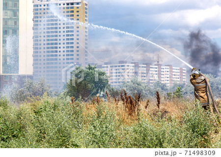 firefighter in a protective suit with a hose pours water over dry grass rescuing from a fire in the background a clearing with dry grass filled with smoke and multi-storey residential buildings. firefighter in a protective suit with a hose pours water over dry grass rescuing from a fire in the background a clearing with dry grass filled with smoke and multi-storey residential buildings. 71498309