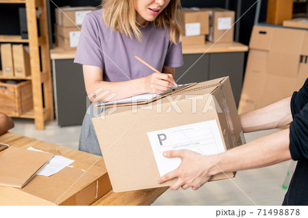 Young female manager of online shop bending over packed box held by man Young female manager of online shop bending over packed box held by man 71498878