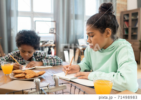 Diligent schoolgirl and her cute brother making notes in copybooks by table Diligent schoolgirl and her cute brother making notes in copybooks by table 71498983
