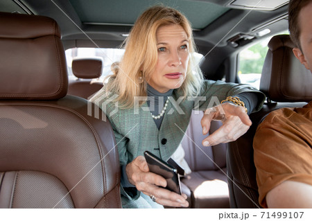 Blond elegant mature businesswoman talking to taxi driver from backseat Blond elegant mature businesswoman talking to taxi driver from backseat 71499107