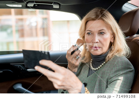 Mature blond businesswoman applying blush on cheeks and looking in small mirror 71499108