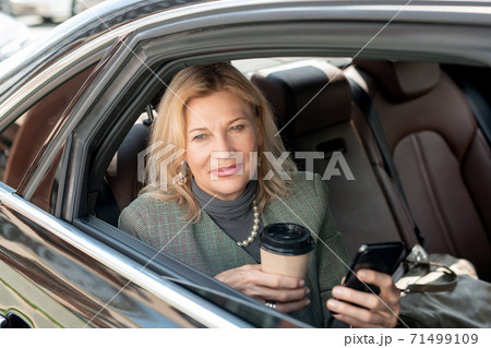Mature woman with glass of coffee using phone while sitting on backseat of car Mature woman with glass of coffee using phone while sitting on backseat of car 71499109