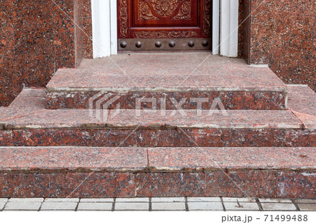 brown wooden entrance door with wood carving pattern on a stone threshold and steps at the granite facade of the building, front view close up. 71499488