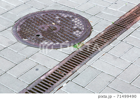 A rust lattice of a drainage paving system and round sewer manhole on a area made of square stone tiles, close up of a rainwater drainage system. 71499490