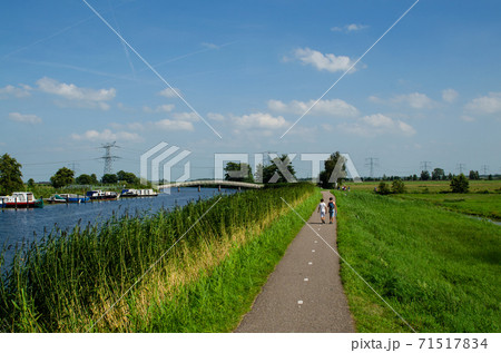Kinderdijk, The Netherlands, August 2019. A small road winds through the Dutch countryside, lined with green grass and a canal, some boys are walking along it. On the left a white bridge. 71517834