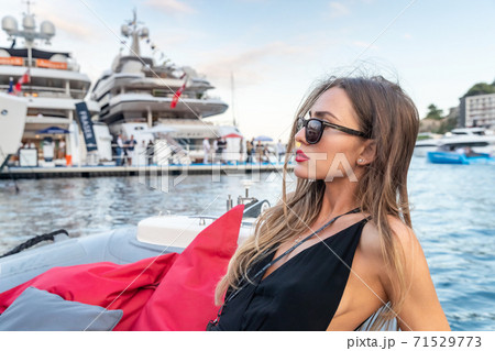 The elegant girl dressed in an evening dress of black color and sunglasses on the boat carry to the big yacht, gorgeous woman, sexual red lips, Decollete, Monaco, Monte-Carlo 71529773