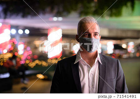 Businessman wearing suit and protective facial mask in city at night while thinking 71541491