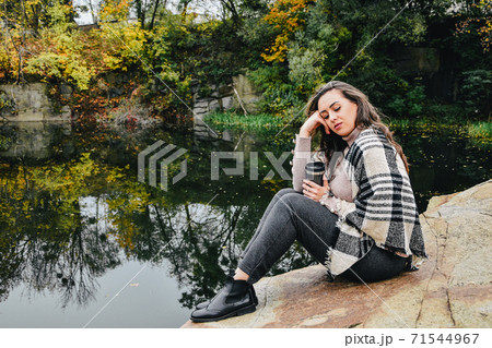 Brunette with long hair sitting next to the lake and drinking hot tea from thermos. Ouutdoor autumn resting. 71544967