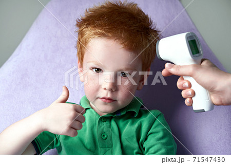 Doctor measures temperature of a cute red haired freckled little boy showing thumb up with a non-contact infrared thermometer. Good health and normal temperature concept. Doctor measures temperature of a cute red haired freckled little boy showing thumb up with a non-contact infrared thermometer. Good health and normal temperature concept. 71547430