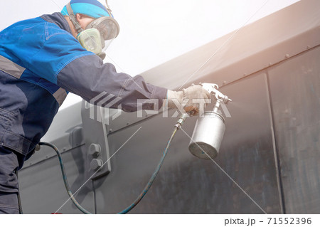 Industrial work. Priming of metal products from the compressor gun. A worker in overalls and a protective mask paints the body of a truck trailer or a metal car. 71552396