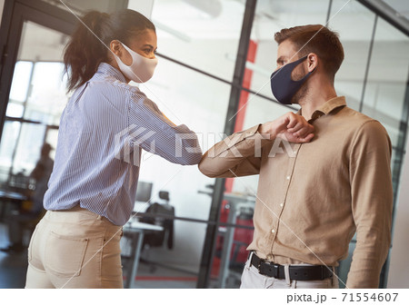 Two young diverse business colleagues wearing face protective masks bumping elbows, greeting each other while working during covid 19 quarantine 71554607