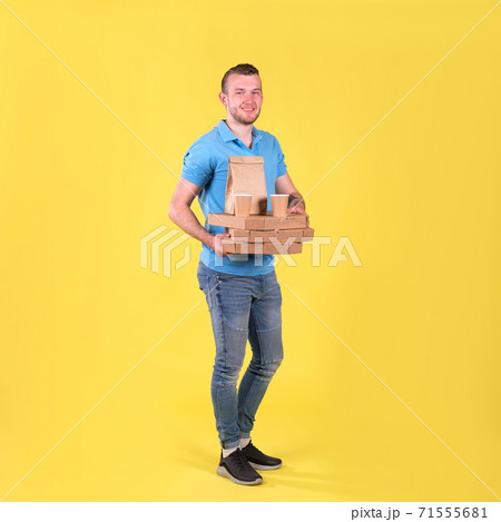 Young smiling guy delivering food from restaurant in blue uniform with thermo bag on his back holds an order for customer on yellow studio background. Fast food delivery to office or home 71555681
