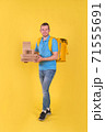 Young smiling guy delivering food from restaurant in blue uniform with thermo bag on his back holds an order for customer on yellow studio background. Fast food delivery to office or home 71555691