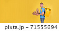 Young smiling guy delivering food from restaurant in blue uniform with thermo bag on his back holds an order for customer on yellow studio background. Fast food delivery to office or home 71555694