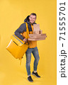 Cute caucasian food delivery courier smiles and looks ahead, holding food order in his hands and yellow refrigerator bag on yellow background hangs on his shoulder. Food delivery to home or office 71555701