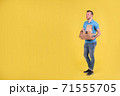 Young smiling guy delivering food from restaurant in blue uniform with thermo bag on his back holds an order for customer on yellow studio background. Fast food delivery to office or home 71555705