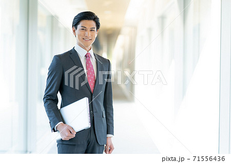 オフィスの廊下でパソコンを持って立つ若い男性のポートレート 71556436