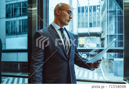 Serious Businessman reads holding paper document. Middle aged Caucasian examines content of business contract standing against of modern building outside the window. Side view 71558098