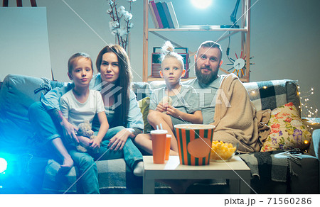 Happy family watching projector, TV, movies with popcorn in the evening at home. Mother, father and kids spending time together. 71560286