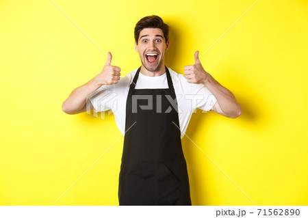 Cheerful man barista in black apron showing thumbs-up, recommending cafe or restaurant, standing against yellow background Cheerful man barista in black apron showing thumbs-up, recommending cafe or restaurant, standing against yellow background 71562890