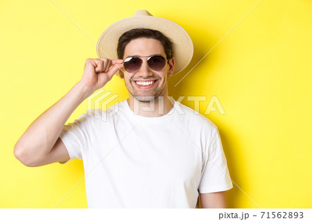 Concept of tourism and vacation. Close-up of handsome man tourist looking happy, wearing sunglasses and summer hat, standing over yellow background Concept of tourism and vacation. Close-up of handsome man tourist looking happy, wearing sunglasses and summer hat, standing over yellow background 71562893