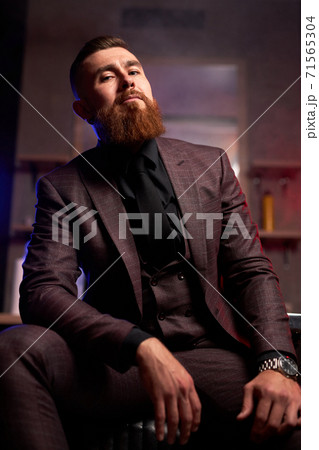 young caucasian handsome bearded man sitting on couch in a dark smoky room 71565304