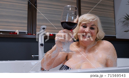 Elderly senior woman lying in warm bath with bubbles, enjoying relaxation, drinking red wine 71569543